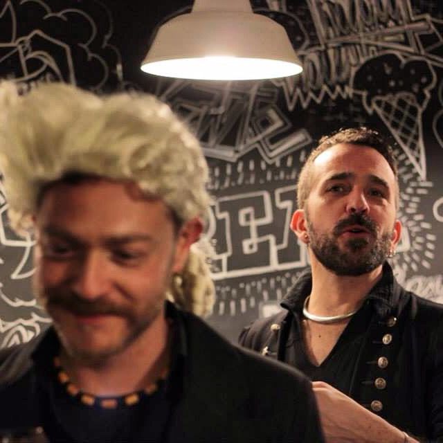 Due attori in costume, uno con parrucca bianca, davanti a un muro disegnato col gesso