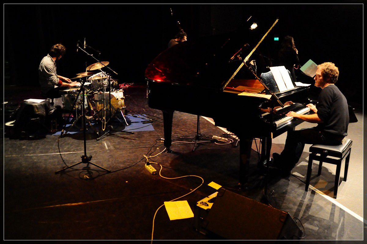 Trio dal vivo su un palco buio: pianoforte a coda, batteria e contrabbasso