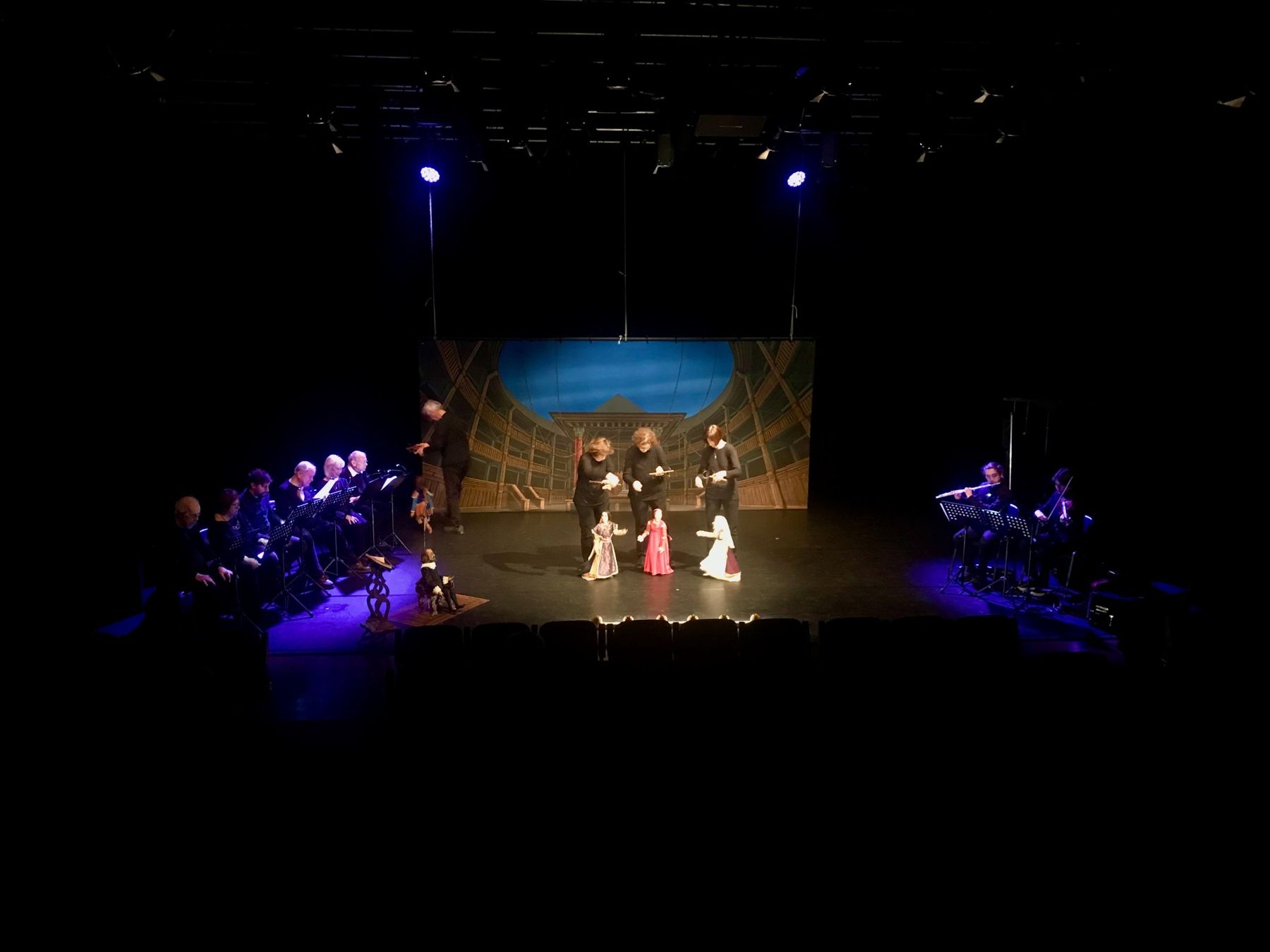 Spettacolo di marionette in teatro, con lettori a sinistra e musicisti a destra della scena