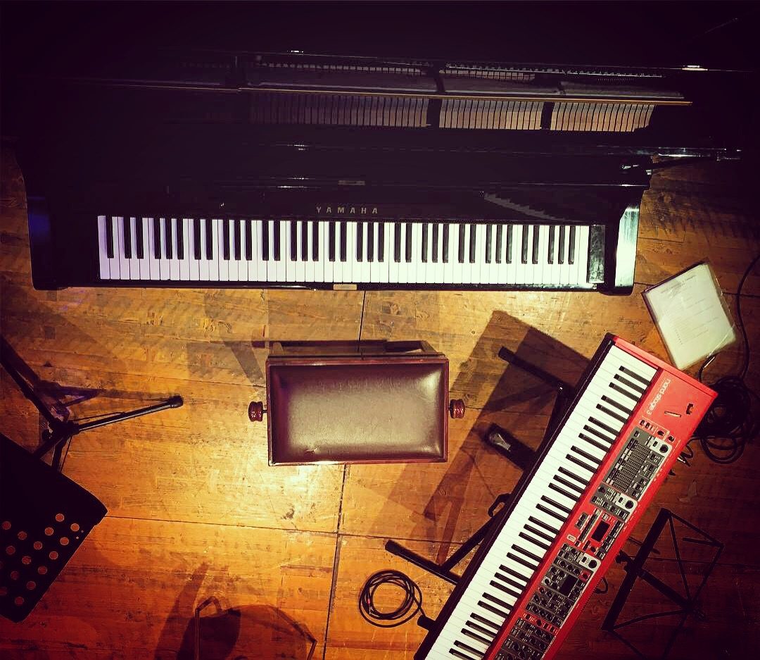 Il palco visto dall'alto: un pianoforte Yamaha, lo sgabello e una tastiera Nord rossa sul pavimento di legno illuminato